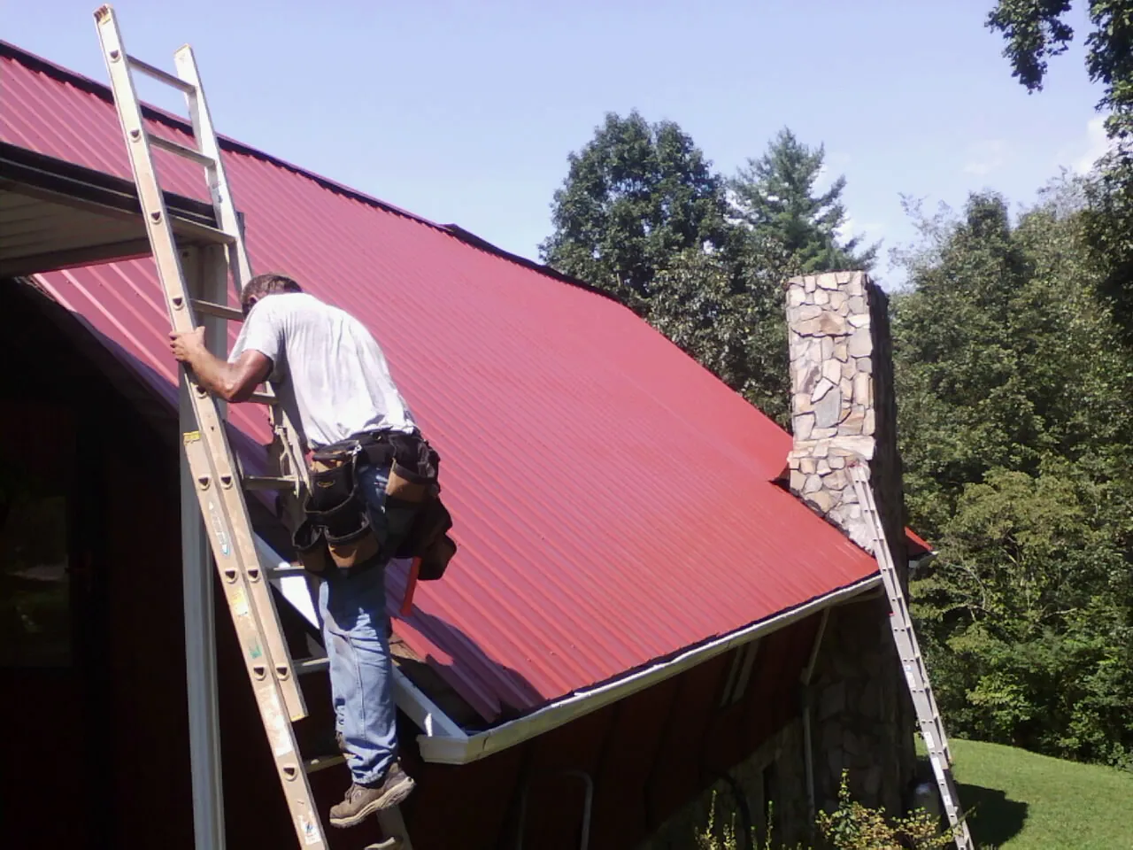 Roofer on a ladder beside a red metal roof and stone chimney — Soffit & Fascia Repair by Roofing Brothers in Seagoville